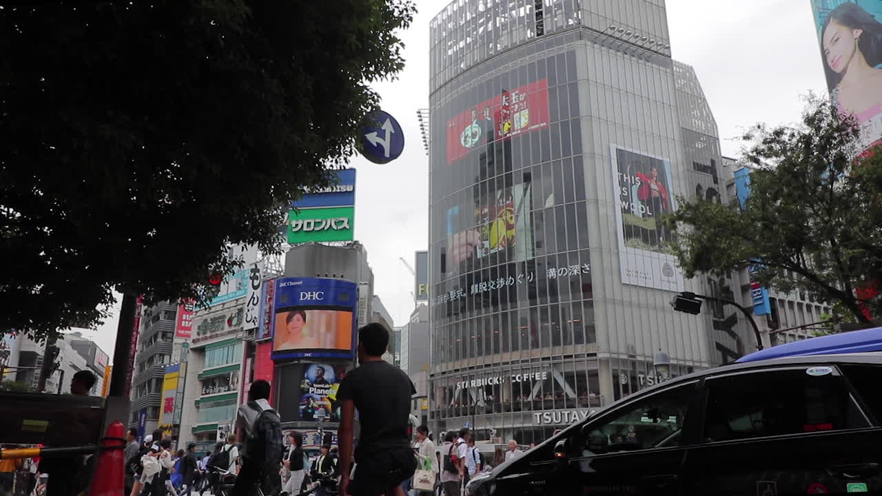 Daytime pedestrians cross the famous busy Shibuya scramble crosswalk in Tokyo while traffic and cyclists wait.