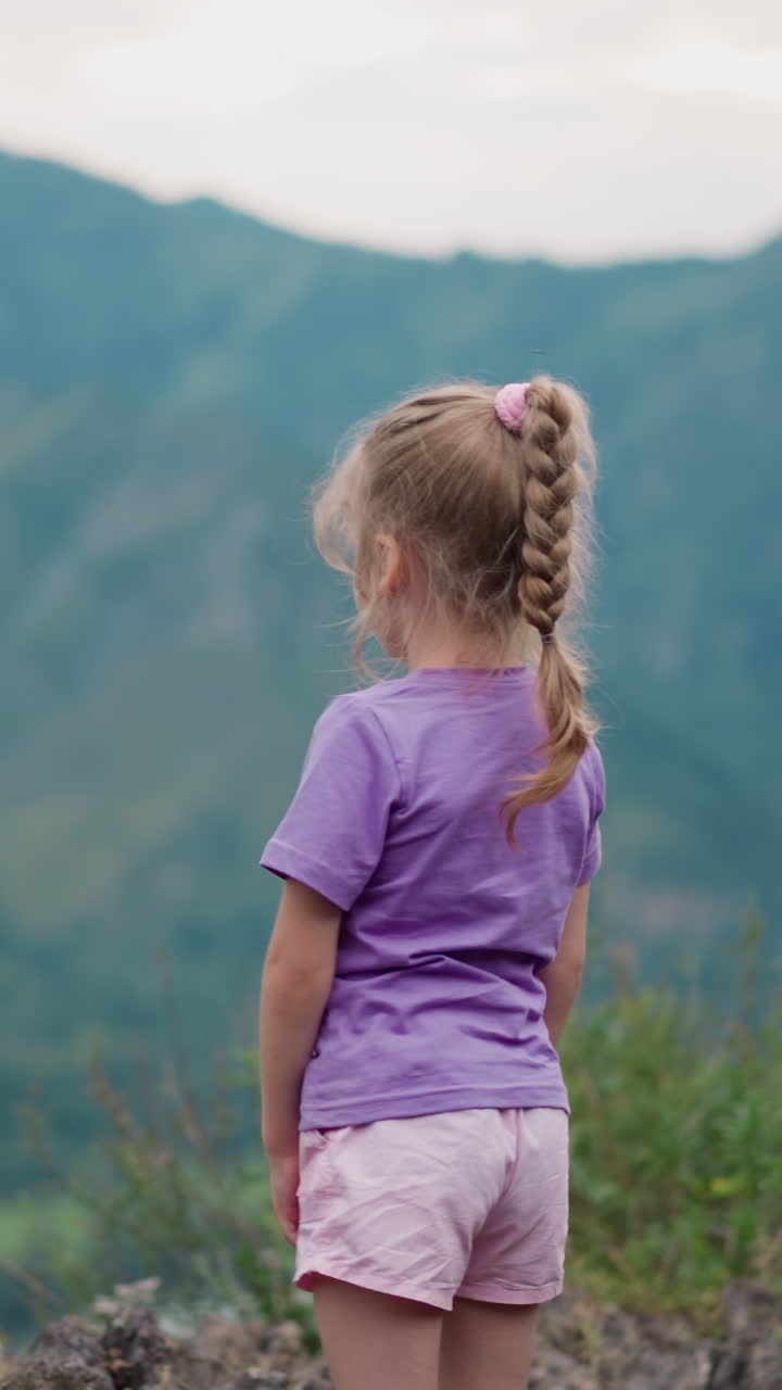 una niña soñadora con una camiseta lila se rasca la pierna con una picadura de mosquito en el borde de la colina contra las montañas en un día sombrío vista trasera en cámara lenta