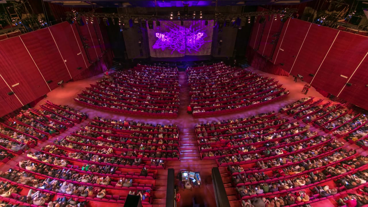 Espectadores se reúnen en el auditorio y ven el espectáculo en el teatro en timelapse. Gran sala con asientos de butacas rojas. Espectadores llenando los lugares hasta que se apaga la luz. Vista desde arriba.