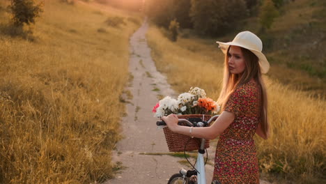 back kind of happy blonde girl in dress and hat turns around and smiling cheerfully looks at the camera and flirts strolling around the field in summer with bike and flowers.