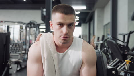 gym trainer sits on weight bench with towel draped over shoulder exhaling in relief after intense chest press workout under bright modern gym lighting showing focused calm expression during recovery