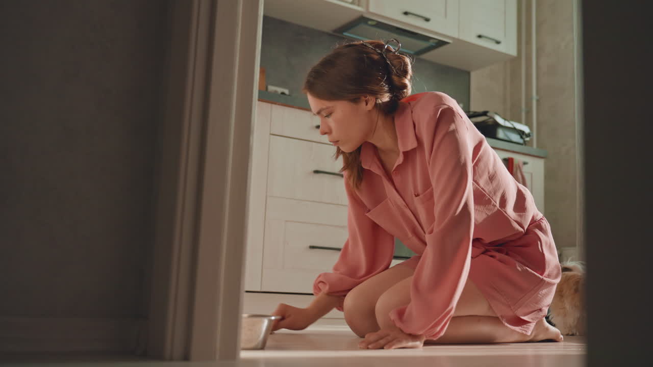 Caucasian Woman Kneeling On Floor Scrubbing Tile With Cloth, Focused Expression, Pink Shirt, Modern Kitchen, Morning Light, Domestic Chore, Routine Maintenance, Practical Determination And Quiet