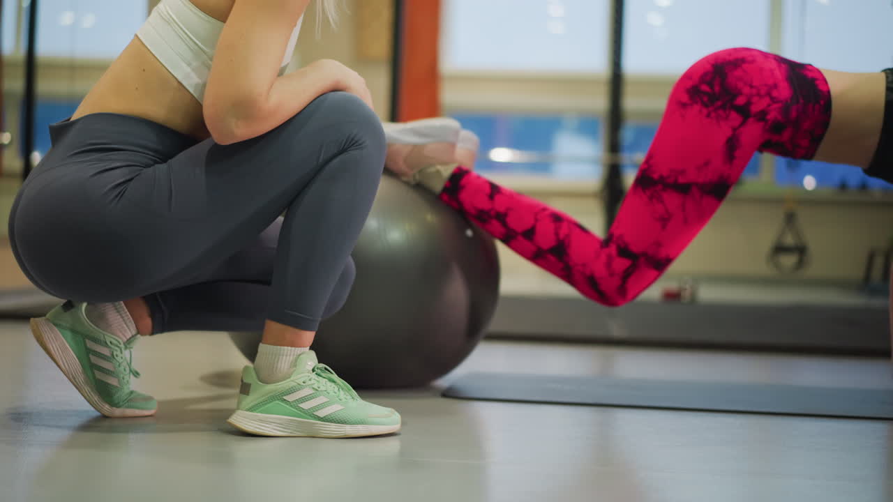 Allenamento intenso del core con palla di stabilità, atleta che esegue flessioni delle ginocchia mentre l'allenatore supervisiona da uno squat, leggings rosa fantasia e sneakers color menta, finestre della palestra e pavimento a specchio, plank dinamico.