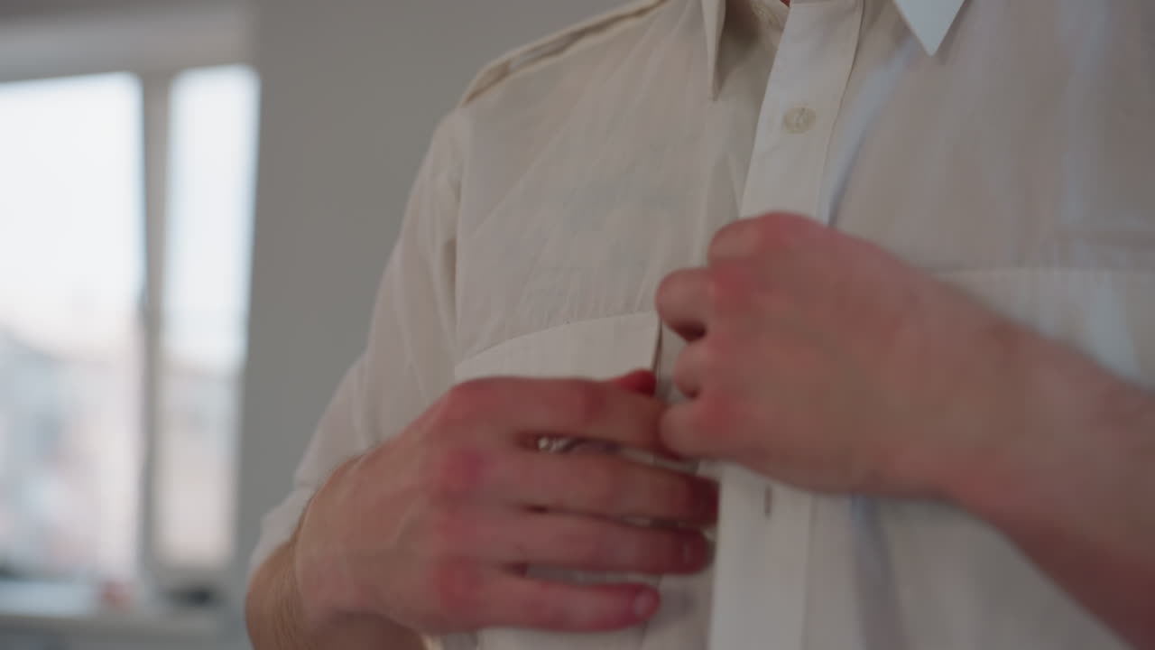 close up of young man carefully unbuttoning white shirt with both hands in daylight room, revealing light fabric and gentle hand movement