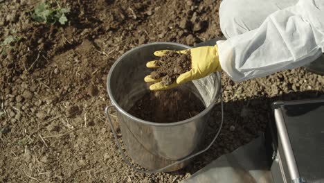 Concepto-De-Muestreo-De-Suelo,-Terreno-De-Campo-En-Manos-Enguantadas-Y-Balde,-Primer-Plano-De-ángulo-Alto