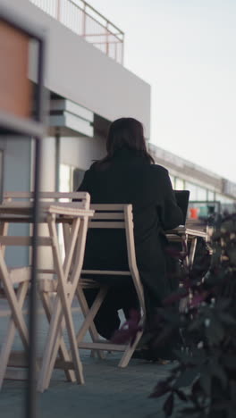 back view of lady in black coat seated at cafe table, typing on laptop, her hair sways gently with the wind in a peaceful urban setting, plants, and wooden chairs around