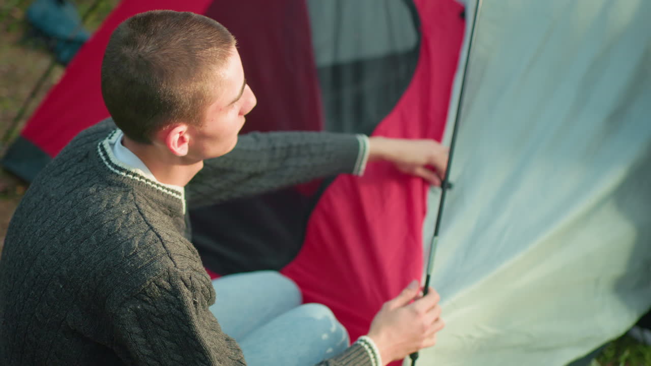 boy kneels beside tent fixing peg to flexible tent pole as sister supports top section during camping in forest sunlight reflects off red and gray tent fabric adding warmth to outdoor teamwork moment