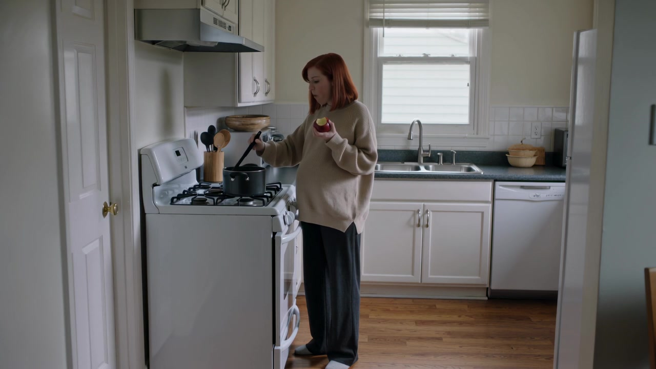 Donna con maglione beige che morde una mela, mescola una pentola e controlla il cibo sul fornello in cucina, con spazio per il testo.