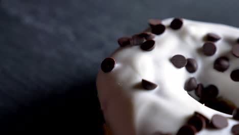 close-up half-shot of glazed white donut with chocolate chips spinning slowly on gray table background.