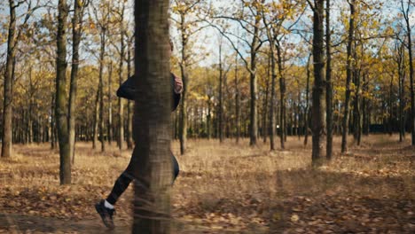 Side-view-of-a-happy-man-with-curly-hair-in-a-black-sports-uniform-running-through-the-morning-autumn-forest-with-fallen-leaves