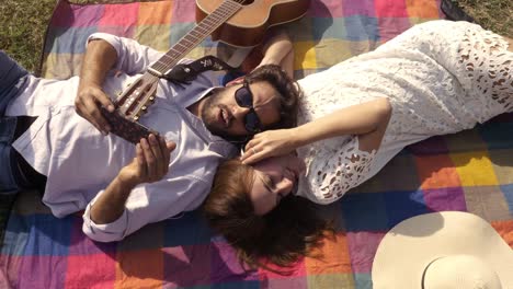 happy lovely young couple lying on colorful blanket in the park listening music on smartphone with hearphones romantic with guitar sunglasses beautiful attractive girl top view rotating camera slow motion