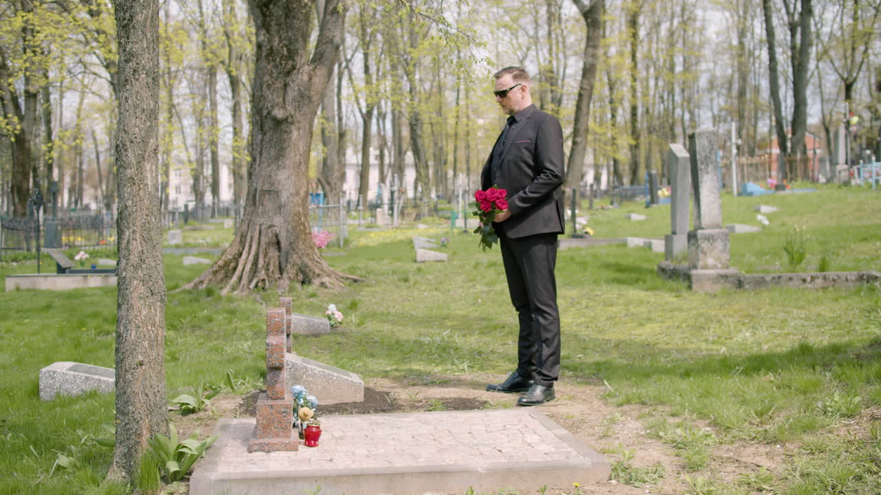 묘지에서 무덤 앞에 서있는 빨간 장미를 들고 검은 양복을 입은 남자