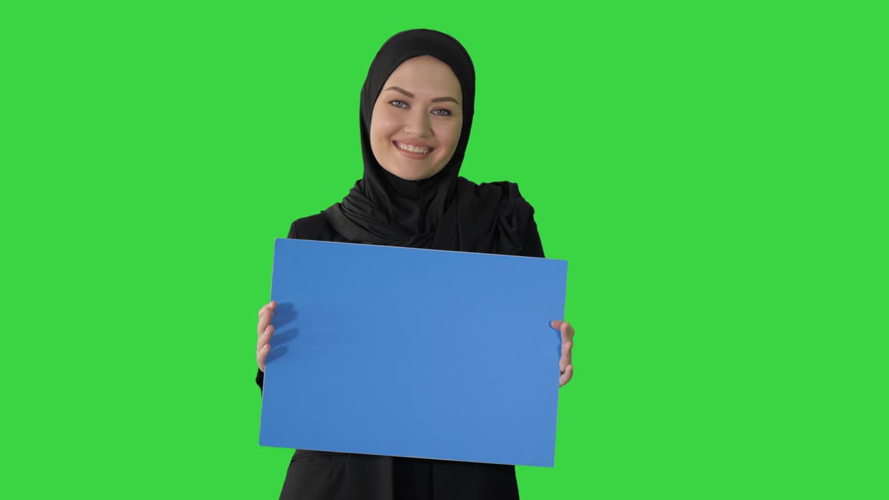 mujer árabe sonriente en hijab sosteniendo un cartel azul en blanco y mirando a la cámara en una pantalla verde, llave de croma