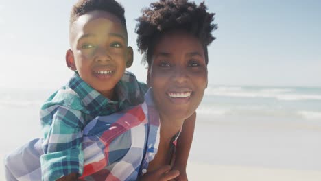 Afroamerikanische-Mutter-Gibt-Ihrem-Sohn-Huckepackfahrt-Am-Strand