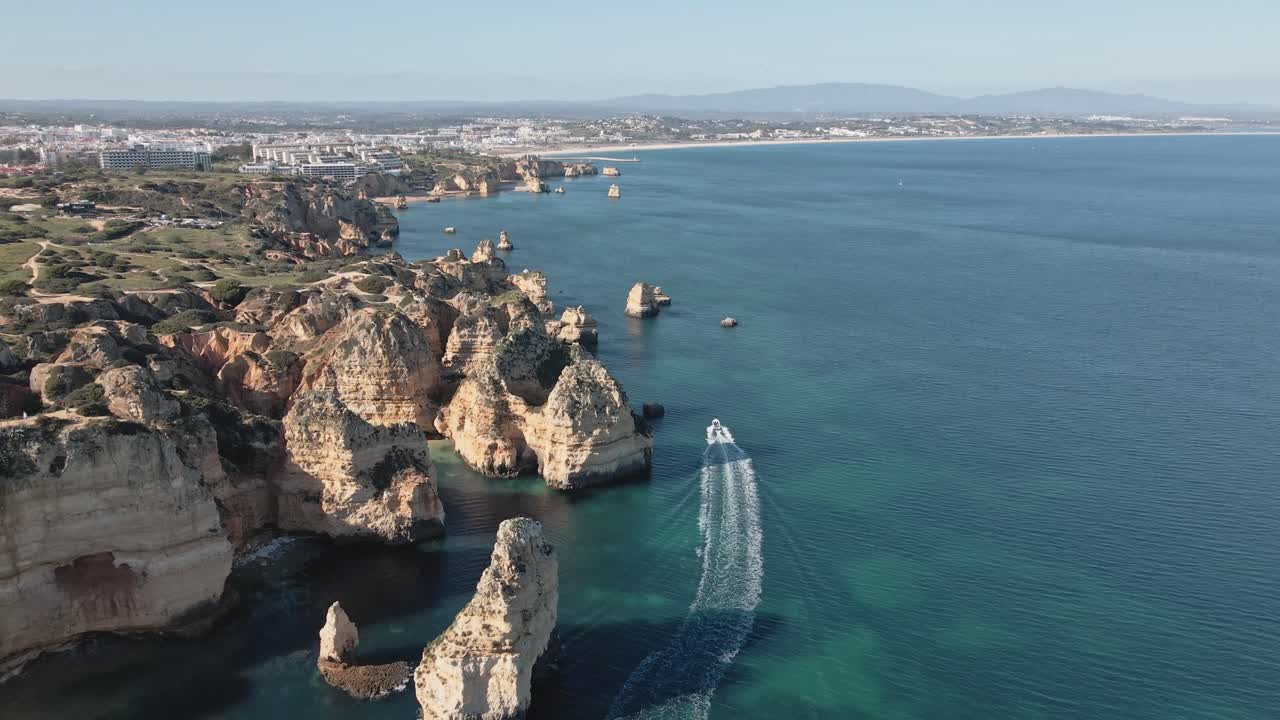 Aerial drone advances above the pristine cliffs and shallow waters of Lagos, Portugal, matching the forward motion of a boat speeding along the turquoise sea