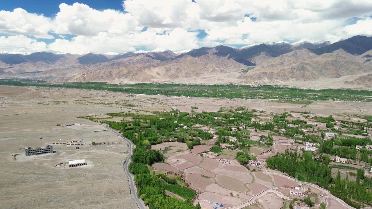 Hindistan'ın Leh Ladakh bölgesinde, bulutlu bir gökyüzü altında, kurak dağ çölü ve engebeli Himalaya sıradağları ile keskin bir tezat oluşturan yemyeşil bir tarım vahası köyünü ortaya çıkaran yüksek açılı drone kaydırma çekimi
