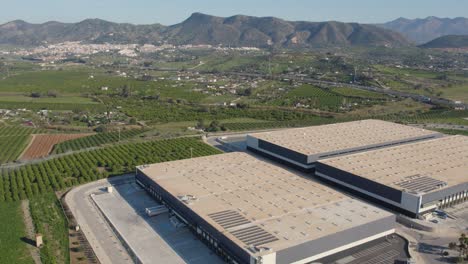 fly-over-a-large-shipping-warehouse-among-the-farmland-of-cartama-spain-on-a-blue-sky-day-with-mountains