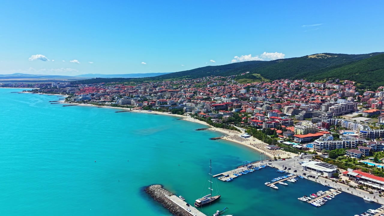 Binalar ve teknelerle dolu, su kenarında bir sahil kasabasının gündüz manzarası, berrak gökyüzü ve masmavi denizle.