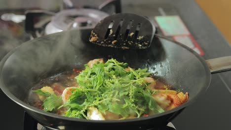 girl is adding fresh bunch of green basil leaves to frying shrimps