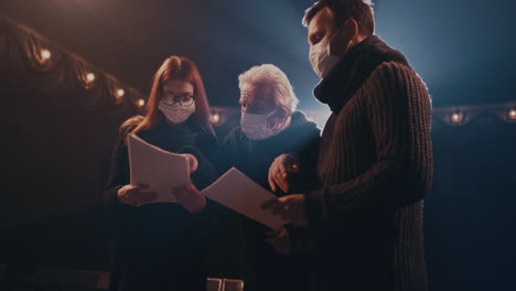 actors in masks discussing script on stage young woman in mask greeting male colleagues with elbow bump then starting to discuss script during rehearsal on stage in theater