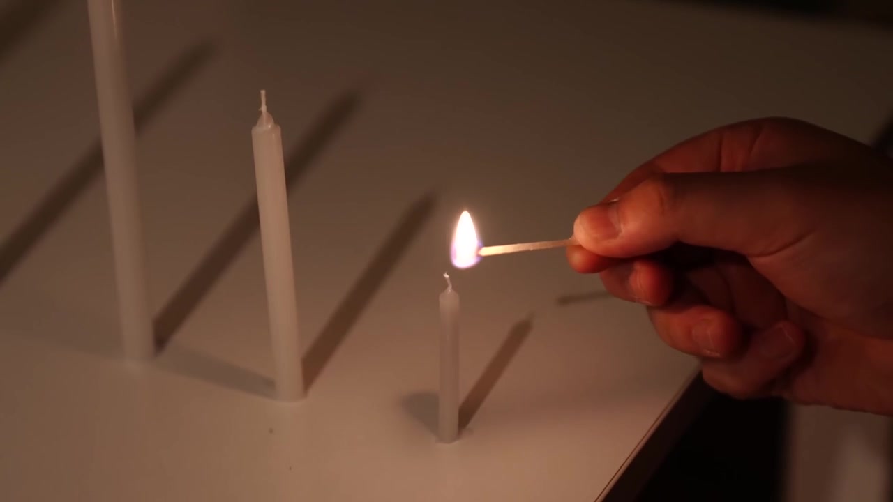 Un ouvrier allumant une petite bougie avec une allumette à côté d'une bougie plus grande non allumée sur une table.
