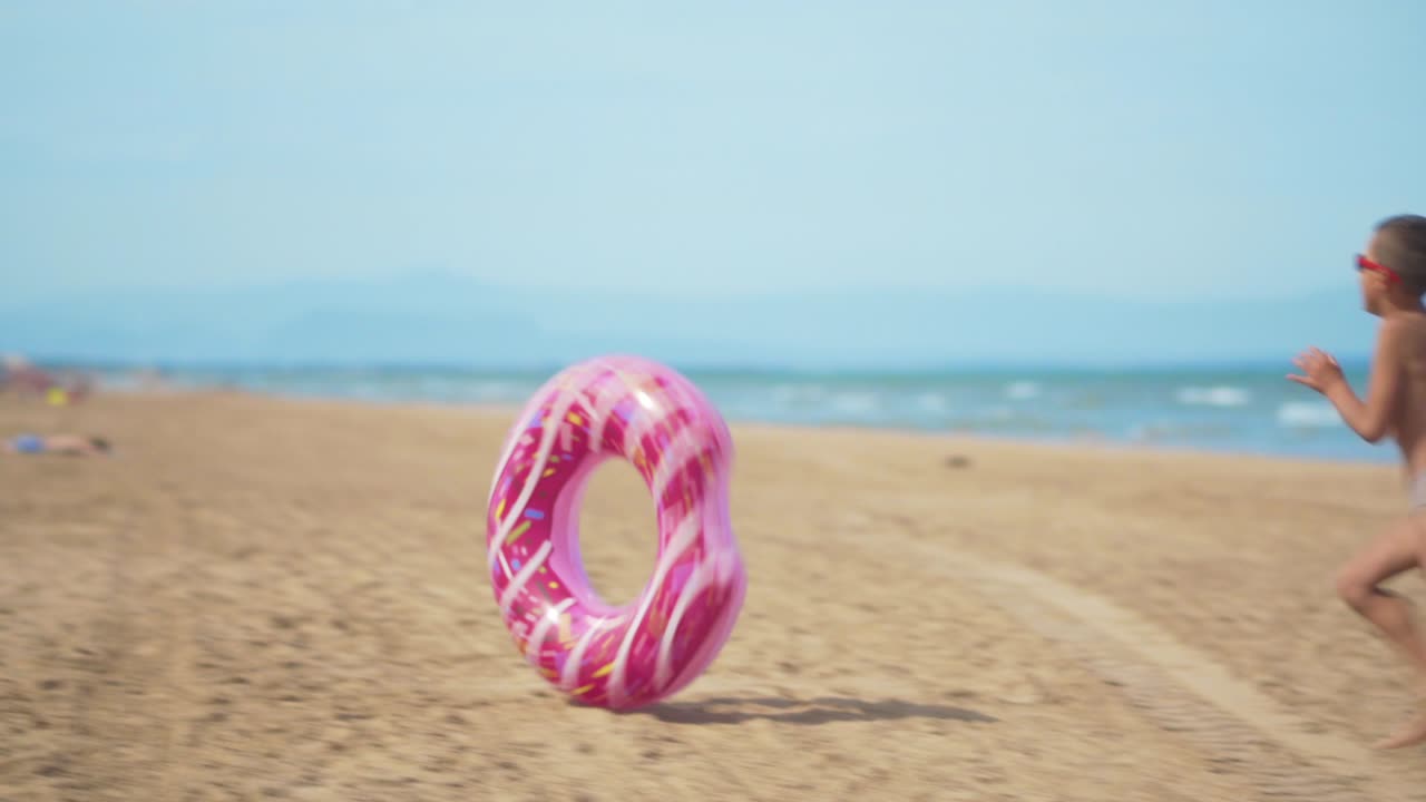 The boy runs along the beach with a pink inflatable donut, rolls it along the sand against the background of the sea. The concept of relaxation and fun