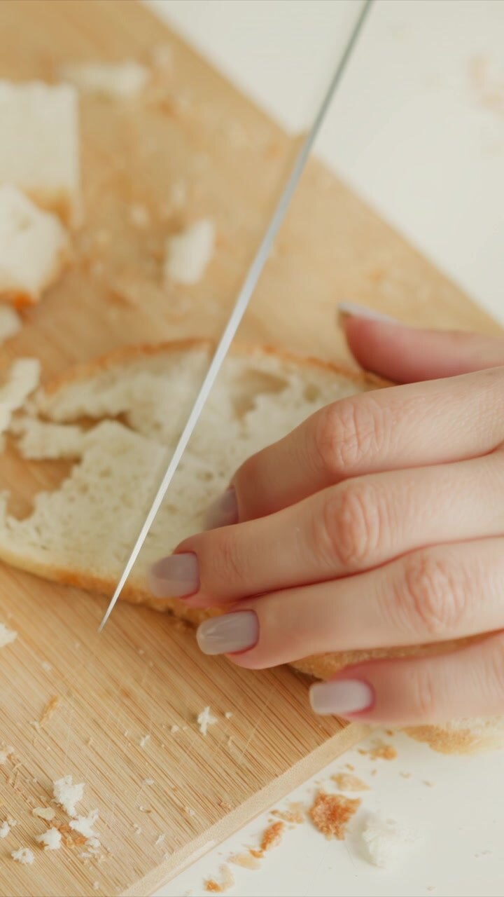 Mano di donna con unghie chiare affetta con cura una pagnotta di pane su un tagliere di legno, creando pezzi uniformi. La vista ravvicinata evidenzia la consistenza del pane bianco fresco e le briciole sparse.
