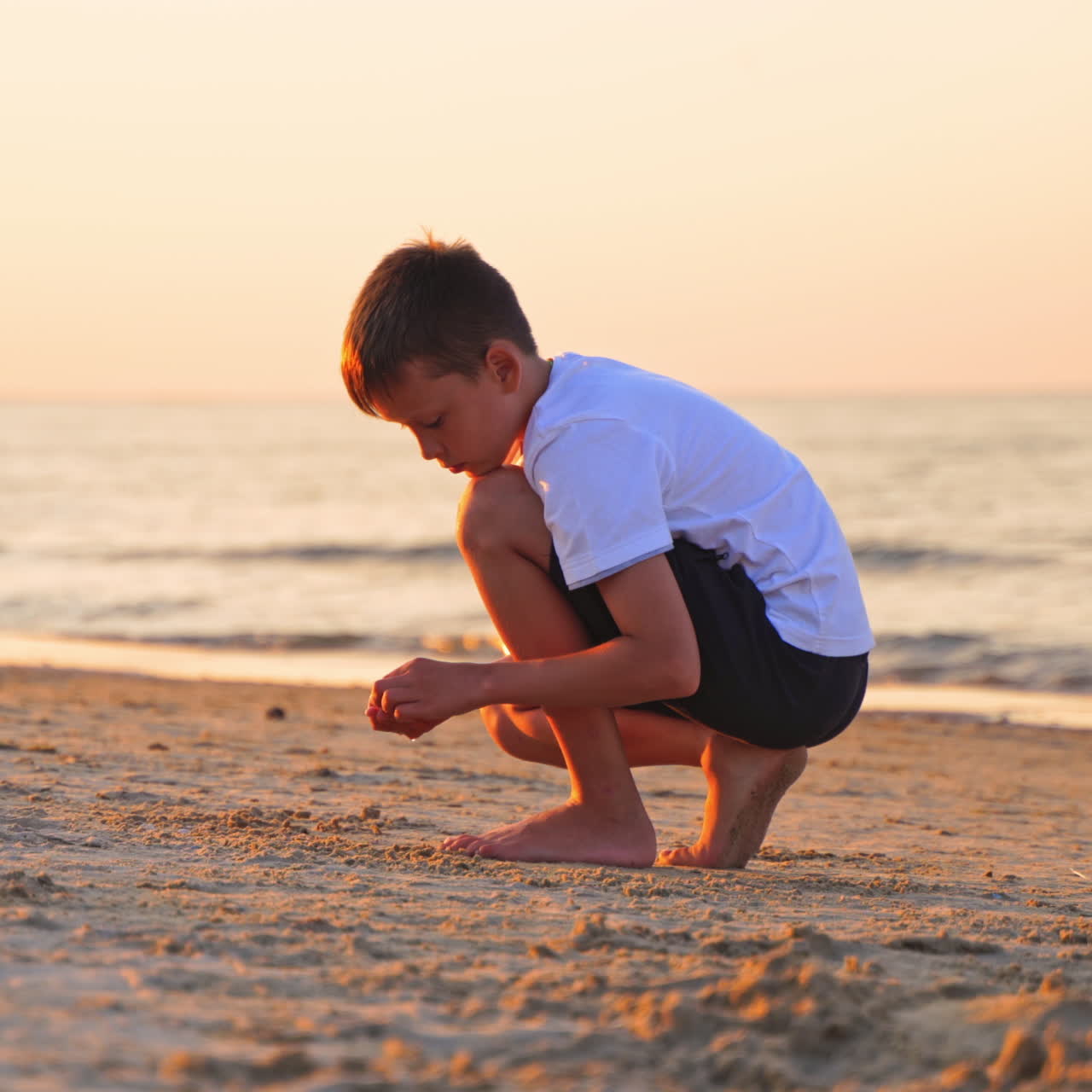 Quiet boy plays on sand beach on sea water background. Evening sea landscape and a young boy putting small pebbles on sand.