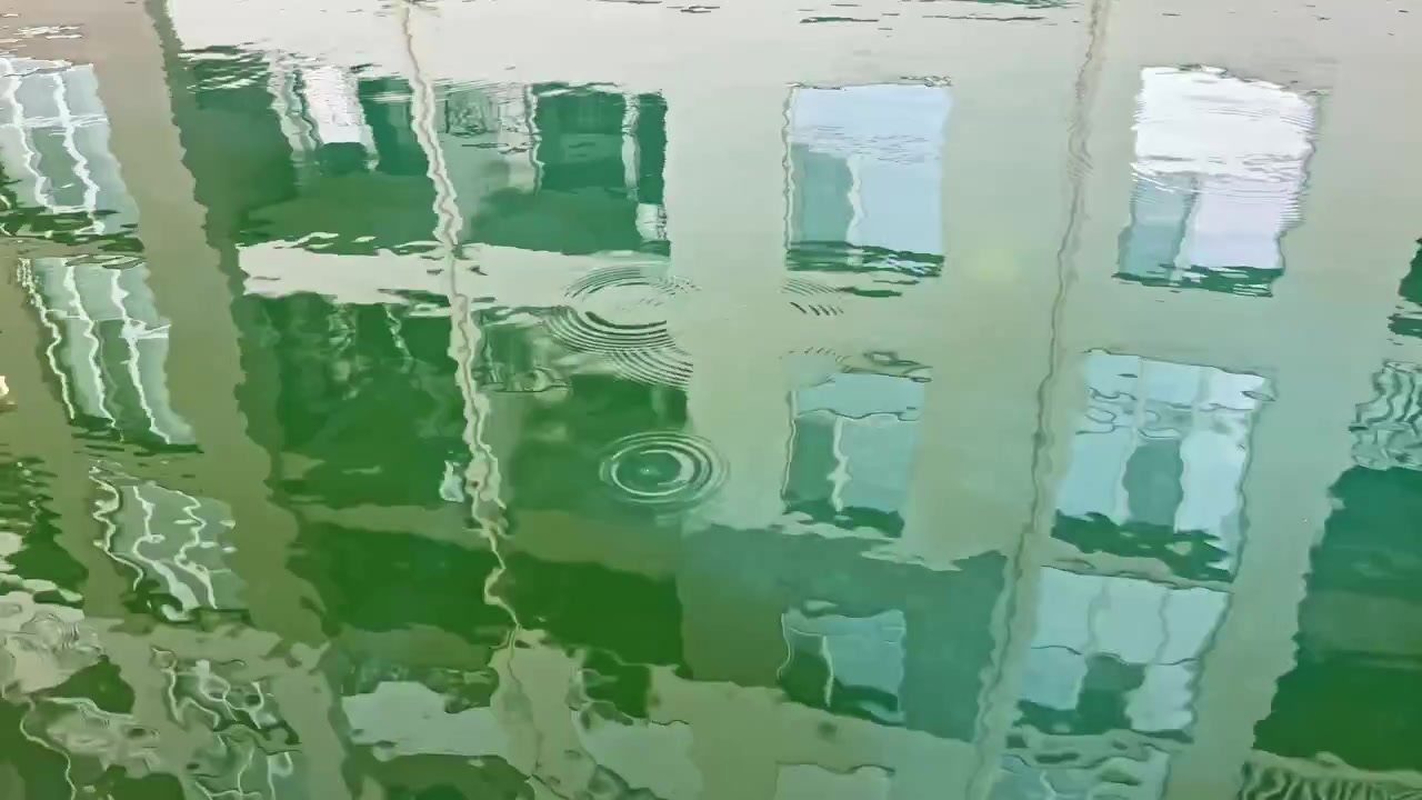 Pantulan bangunan di atas air hijau di bawah cahaya siang yang terang dekat kanal dengan riak-riak lembut dan bayangan halus di pemandangan.