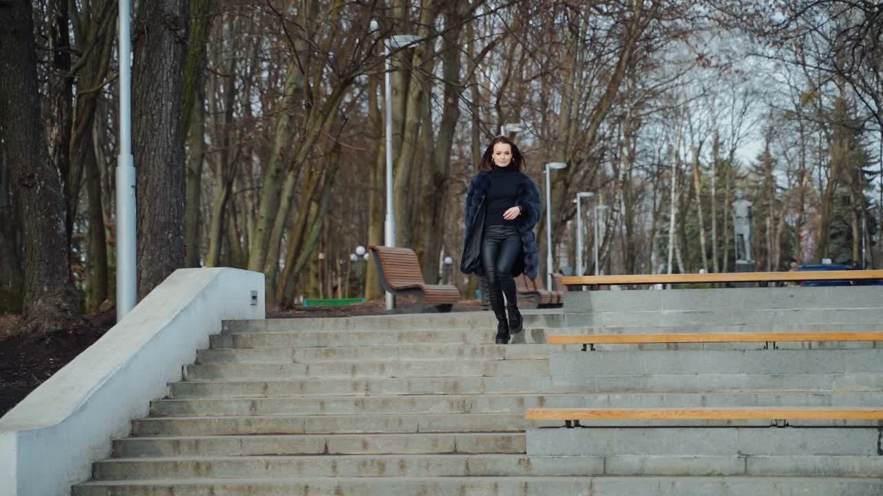 Beautiful girl in the park. Happy young woman goes down the stairs in the park. Attractive model in fur coat. Slow motion.