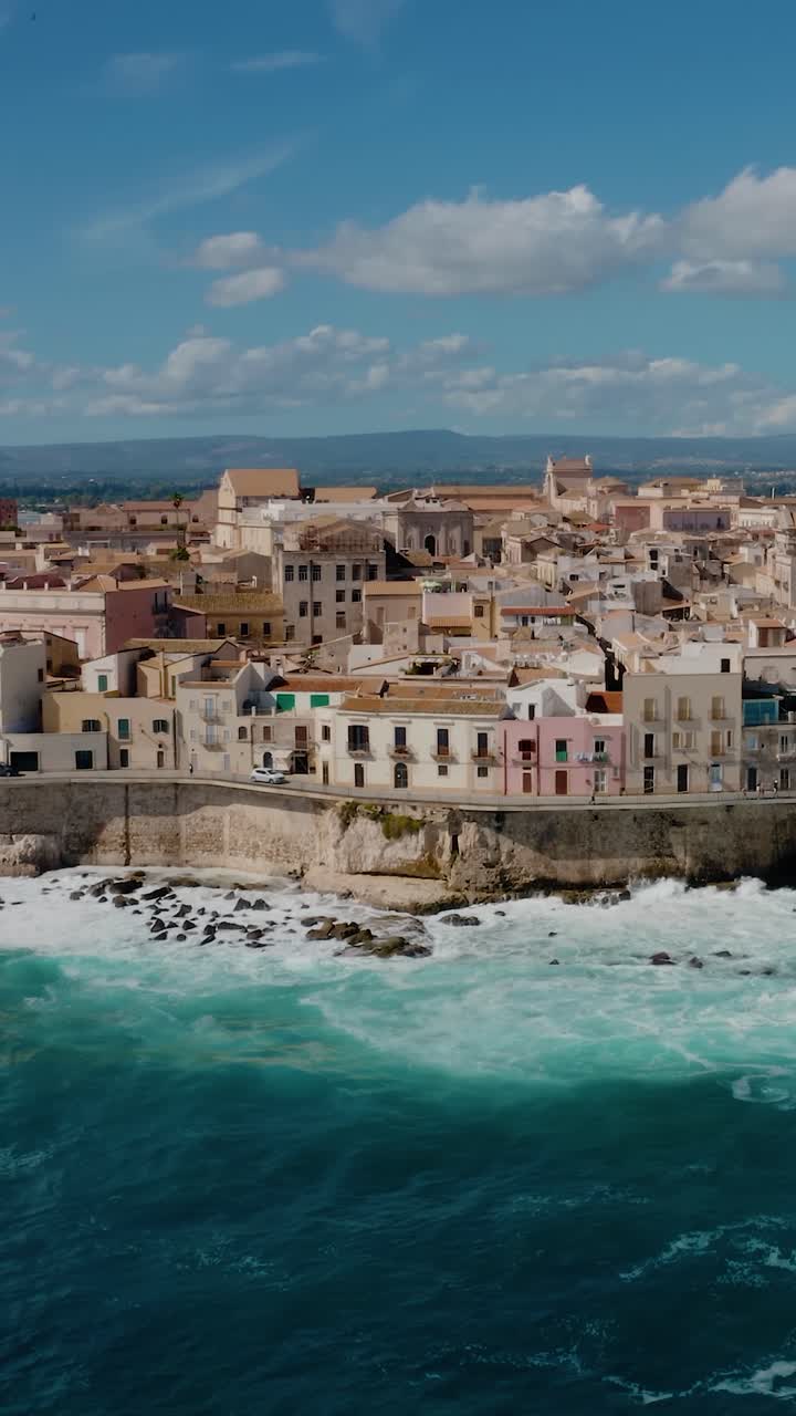 Vertical aerial drone shot of Ortigia island in Syracuse. Historic architecture on a natural fortress by Mediterranean sea with waves crashing on the coast. UNESCO world heritage site in Sicily.