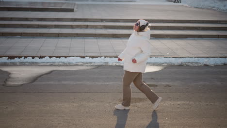 young woman glides and twirls along snow lined city sidewalk crossing street with arms raised wearing puffy coat beanie headphones and sunglasses moving to rhythm under winter sun glow