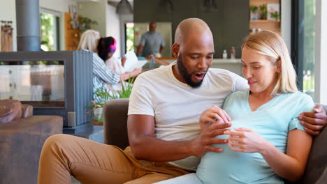 Multi-Racial-Couple-With-Pregnant-Woman-On-Sofa-At-Home-With-Multi-Generation-Family-In-Background