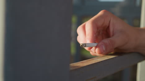close up of fair skinned person casually holding cigarette over metal surface while wind blows ash and fingers slowly roll cigarette under soft daylight in relaxed urban setting