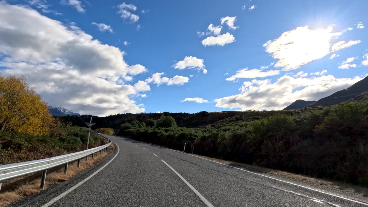 Vehicle travels winding mountain road, bright daylight, dramatic clouds, lush landscape, Queenstown, New Zealand