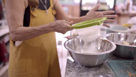 Mujer-Madura-Tamizando-Harina-En-Un-Recipiente-De-Metal-En-La-Cocina