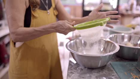 mujer madura tamizando harina en un cuenco de metal en la cocina
