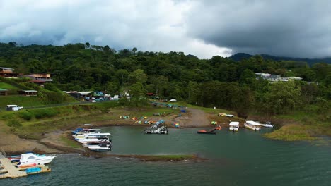 reverse aerial pan retreats across moored fishing boats and wooden pier in quiet lakeside harbour of rustic village on lake arenal, costa rica, framed by tropical hills and calm reflective water