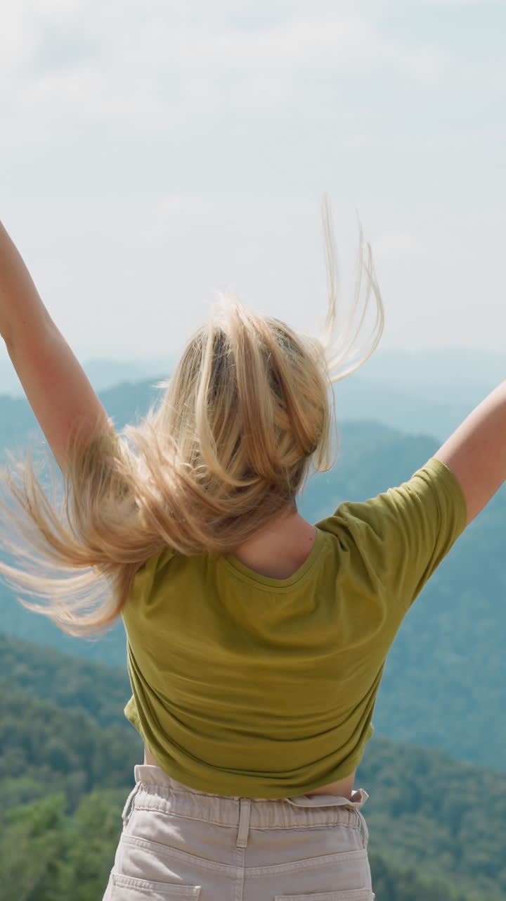 Lady in shorts and t-shirt plays with loose hair and enjoys rejoicing with inspiring valley view against mountains silhouettes in summer backside view