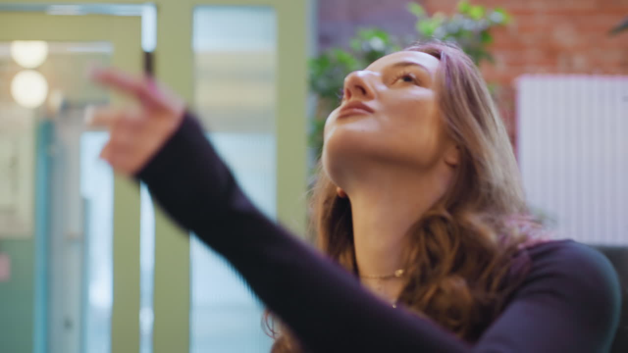 Confident woman with long curly hair and subtle makeup wearing black top and necklace stands calmly while admiring indoor greenery with soft smile in peaceful, light-filled modern space