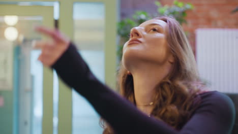 Confident woman with long curly hair and subtle makeup wearing black top and necklace stands calmly while admiring indoor greenery with soft smile in peaceful, light-filled modern space