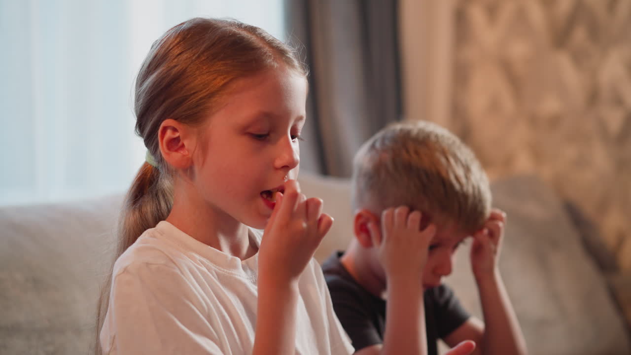 girl child dipping fries into sauce enjoying eating with little boy beside her scratching head while both look curiously at someone in background seated together
