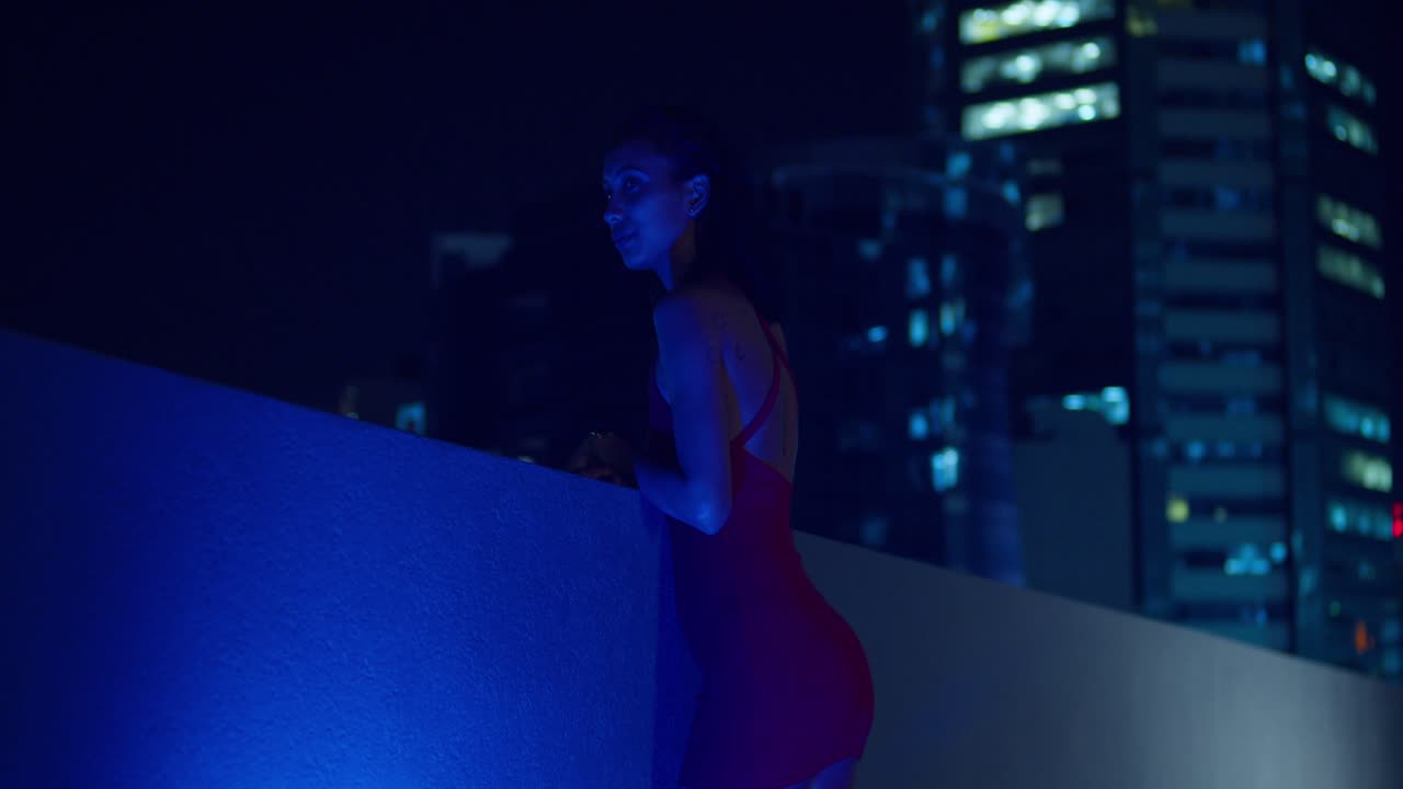 Una ragazza in abito rosso si trova su un tetto di notte, con gli edifici della città sullo sfondo