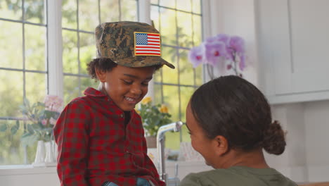 american army mother in uniform home on leave playing with son wearing her cap in family kitchen