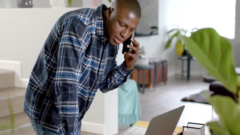 African-american-man-using-laptop-and-talking-on-smartphone-at-home,-slow-motion