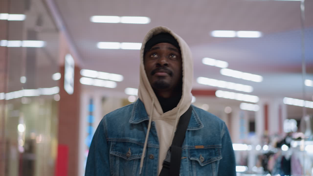 young black man wearing beige hoodie under denim jacket glancing right with subtle smirk while walking through brightly lit shopping mall corridor with blurred displays