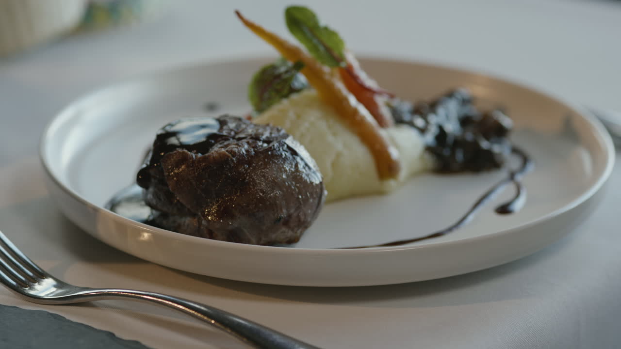 Close-up of a waiter serving a gourmet dish with mashed potatoes, baby carrots, sprouts, and a piece of meat with dark gravy