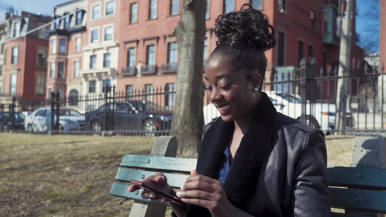 Attractive Young African American Woman Using Smartphone Sitting On Park Bench, Receiving Text Message That Makes Her Laugh And Smile, Sunny Day In The City