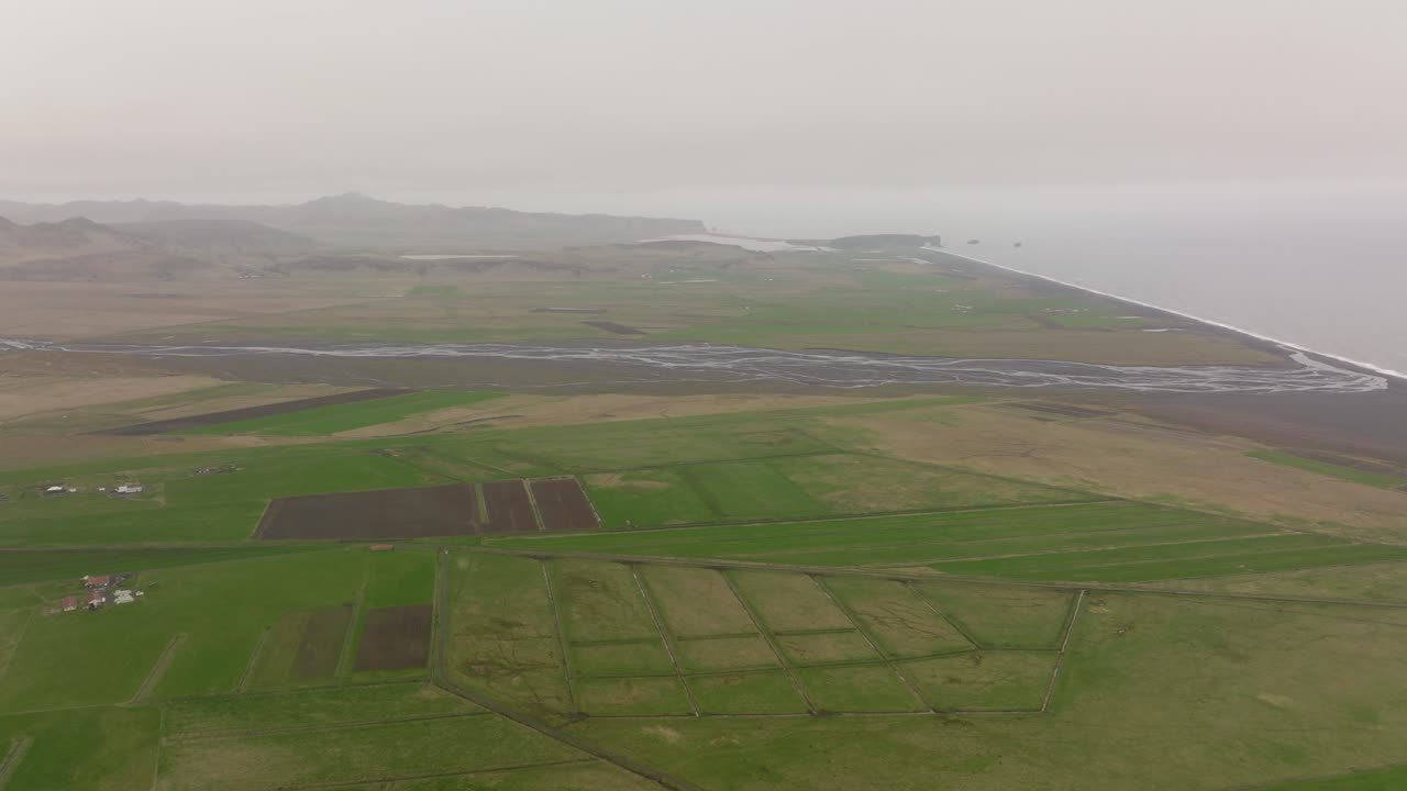 Aerial shot of the farmlands and coastal plains near Petursey, Iceland, showcasing a patchwork of green fields and winding rivers flowing toward the ocean.