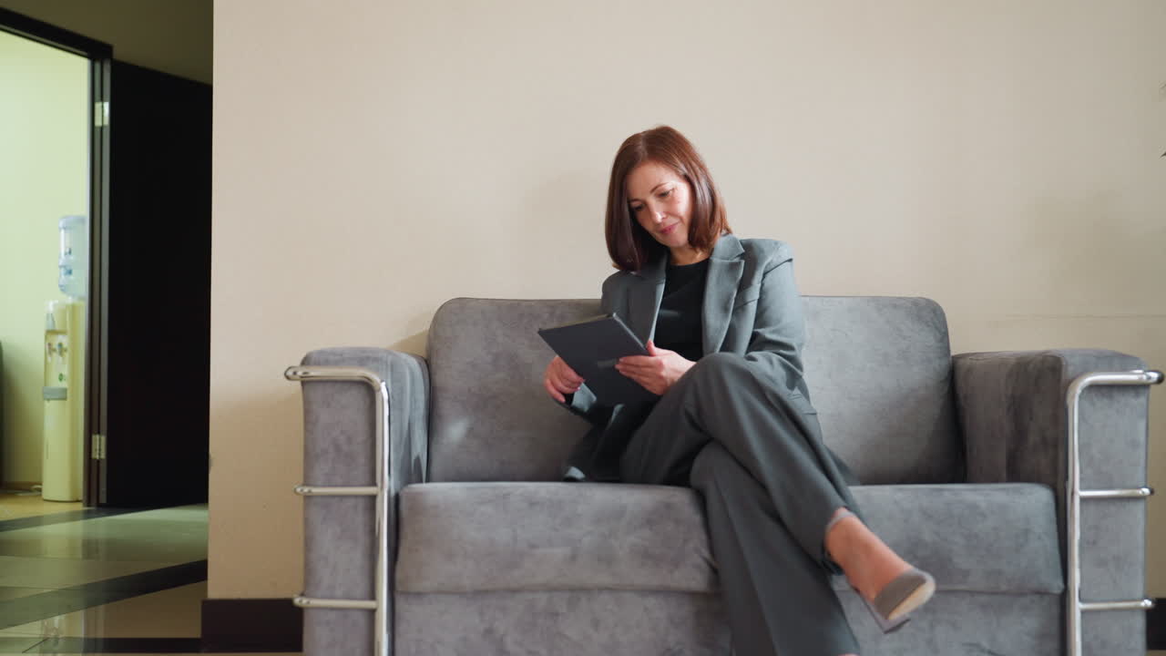 Confident businesswoman in gray pantsuit sitting on modern sofa using tablet in bright office reception area, smiling slightly while working or reading with legs crossed near indoor plant and open doorway